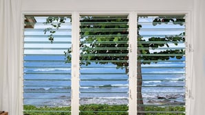A room with a view of the ocean through a window with horizontal blinds.