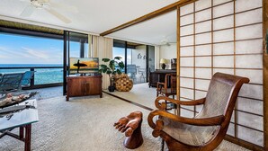 Una habitación amplia con un gran ventanal con vistas al océano, una silla de madera, una mesita y un ventilador de techo.
