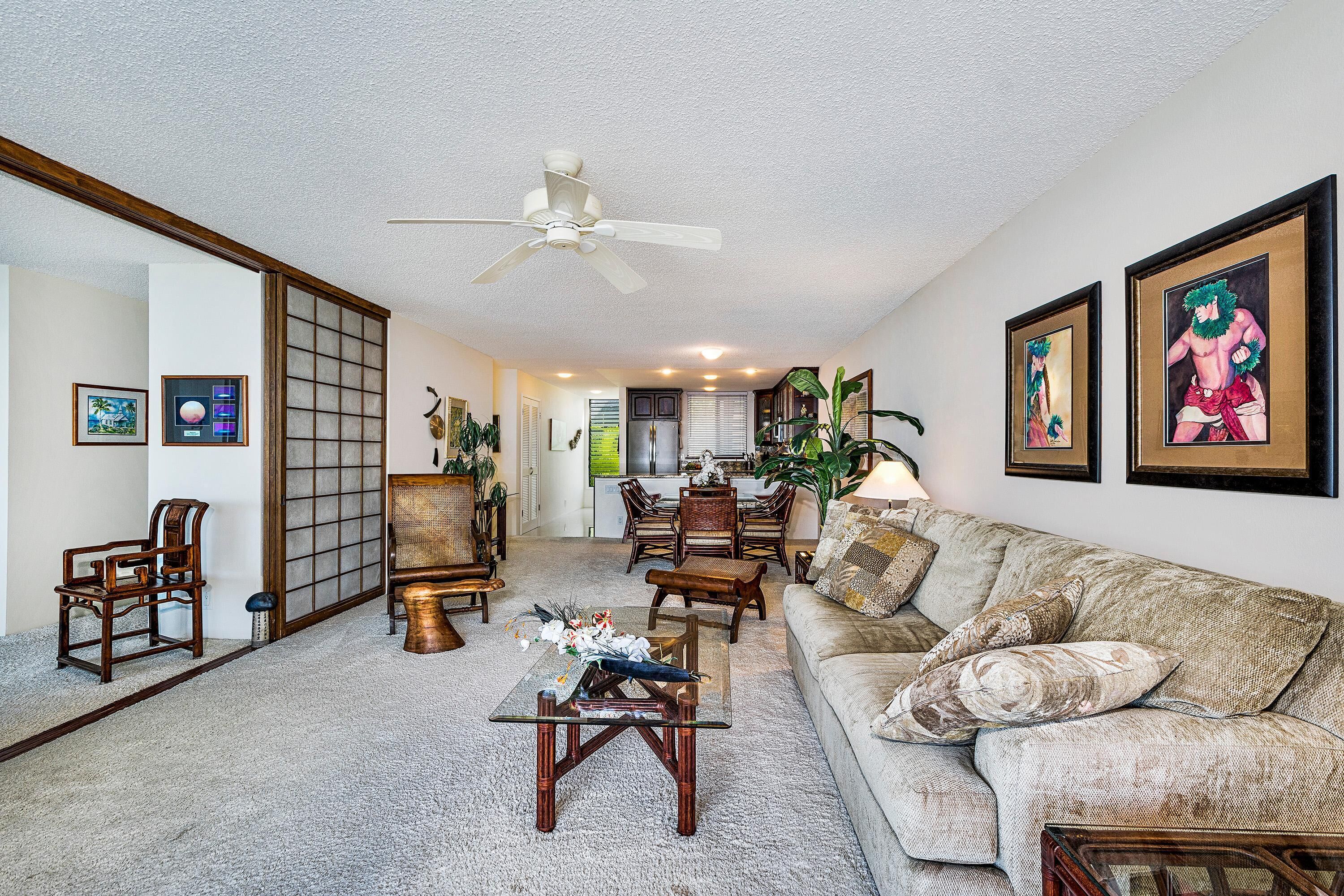A spacious living room with a sofa, coffee table, and chairs. A ceiling fan is visible, and there are framed pictures on the walls.