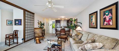 A spacious living room with a sofa, coffee table, and chairs. A ceiling fan is visible, and there are framed pictures on the walls.