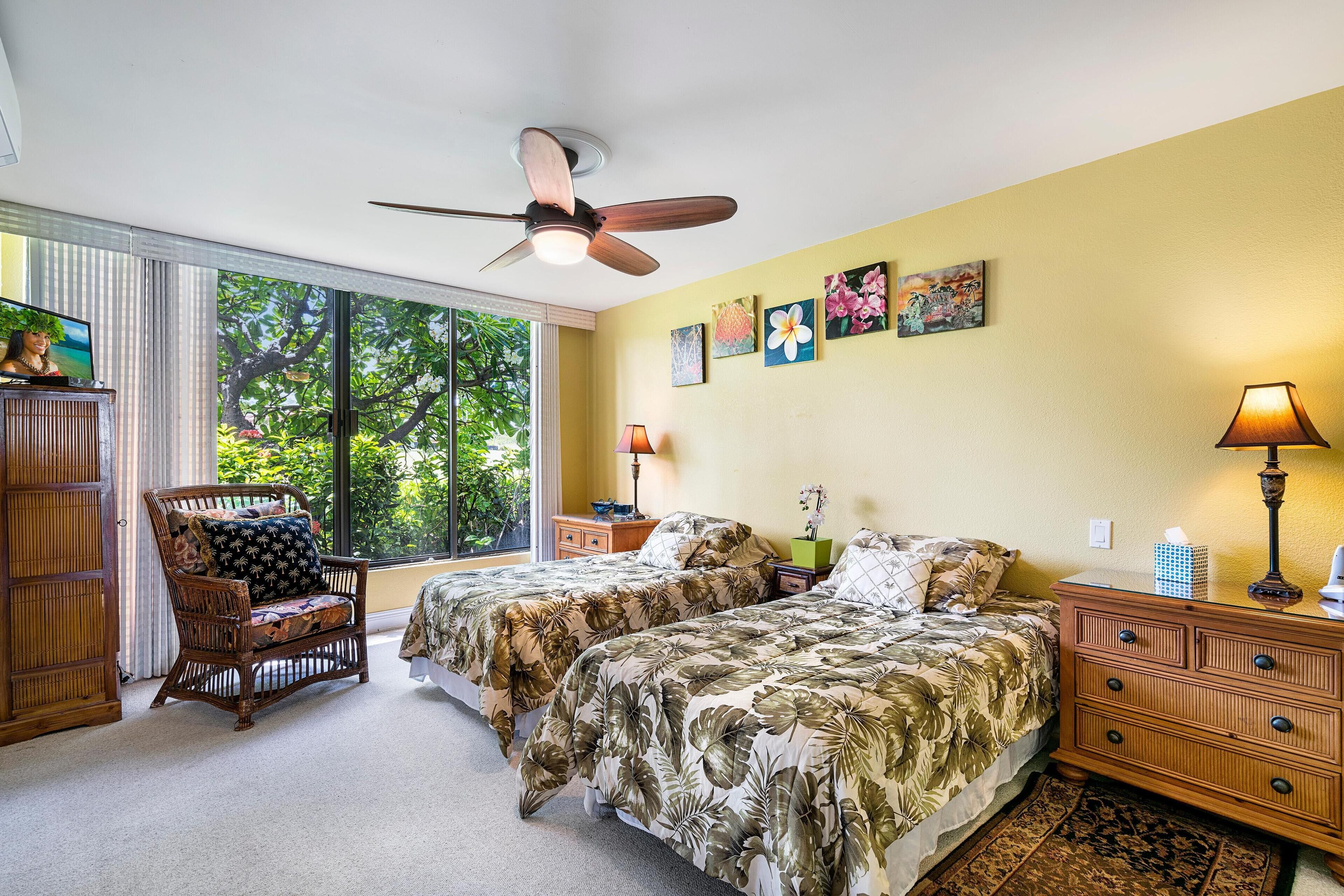 A bedroom with two beds, a ceiling fan, a window with a view of trees, and artwork on the wall.