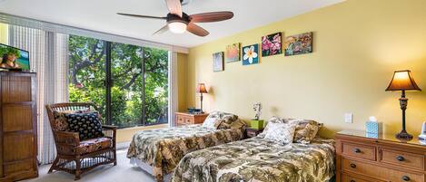 A bedroom with two beds, a ceiling fan, a window with a view of trees, and artwork on the wall.
