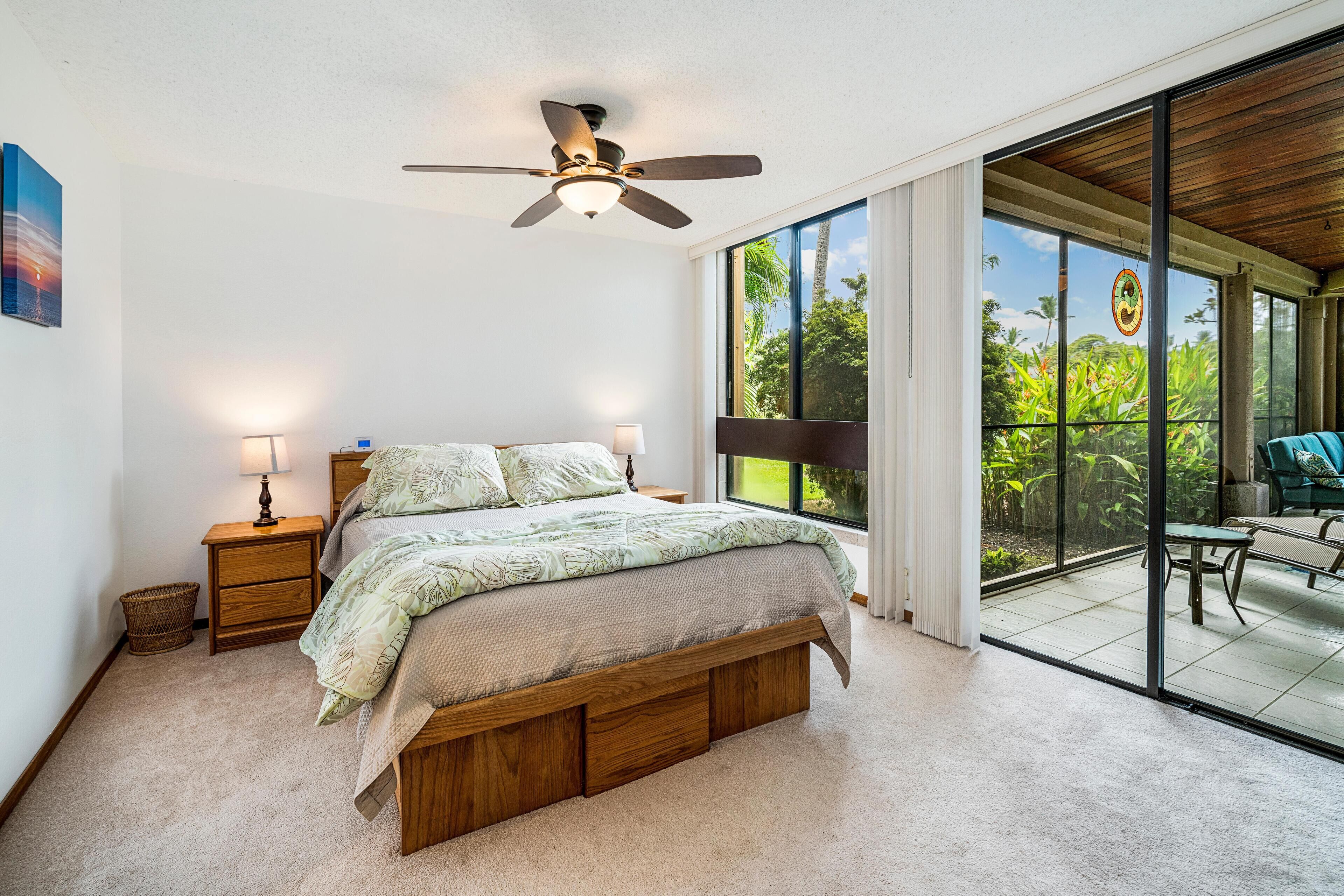 A bedroom with a large bed, wooden headboard, two bedside tables with lamps, a ceiling fan, and a view of a patio with outdoor furniture.