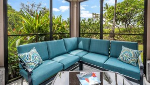A teal sectional sofa with patterned cushions, a glass coffee table, and a view of lush greenery and trees through large windows. - Keauhou Kona Surf & Racquet Club #6-104 (Kailua-Kona)