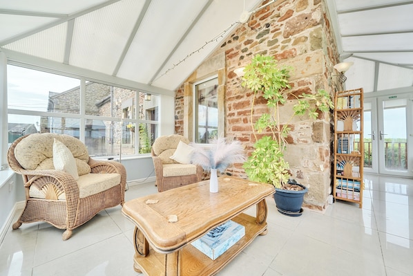 A spacious room with a high ceiling, stone walls, and large windows. There are wicker chairs and a wooden table with a chess set.