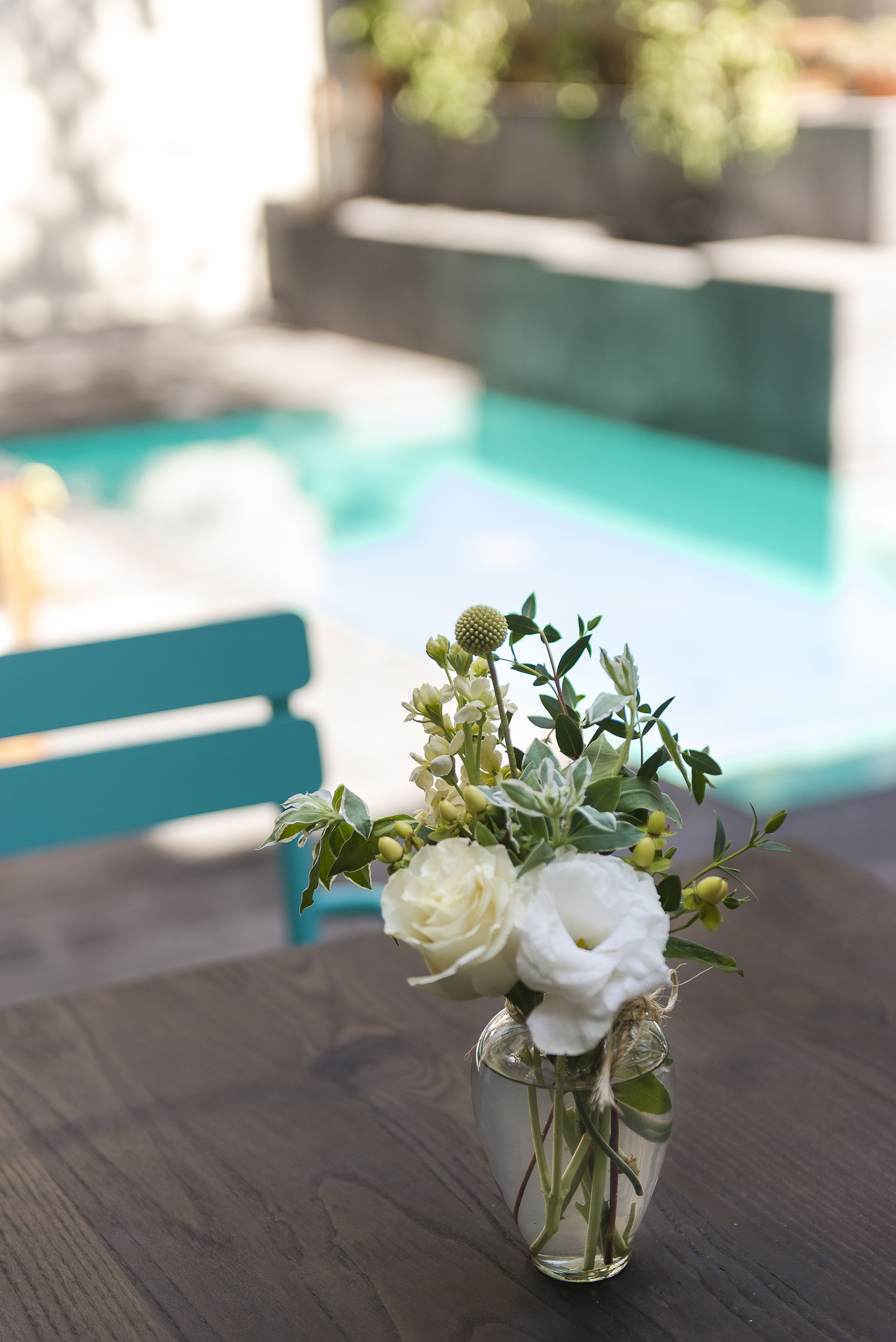 A clear glass vase with white flowers and green foliage on a wooden table by a pool.