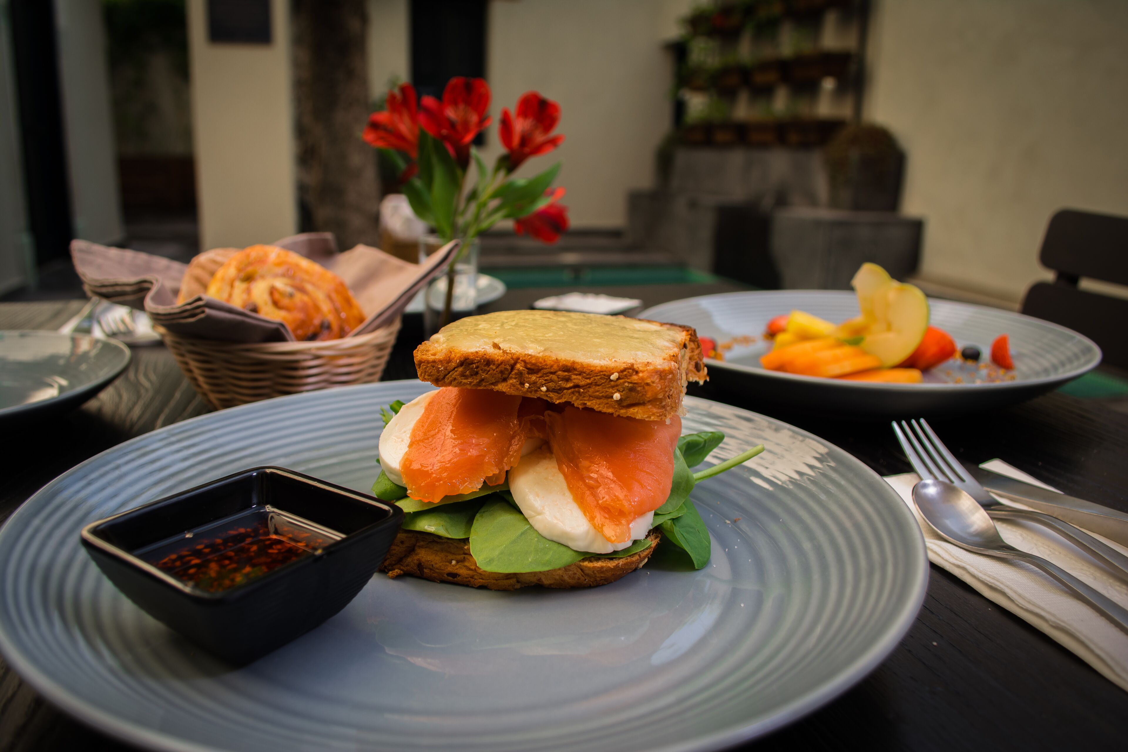 A sandwich on a plate with a side of dipping sauce, a basket of bread, and a plate with fruit.