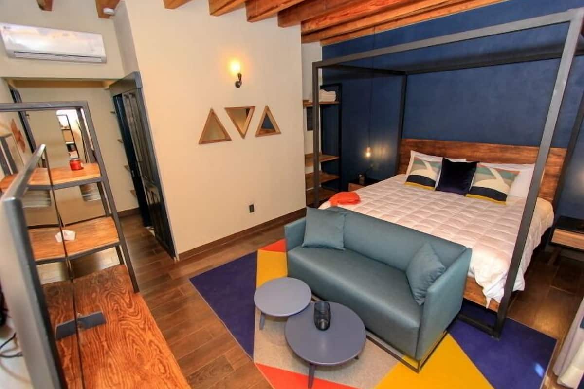 A modern hotel room with a bed, a sofa, and a small table. There is a wooden ceiling and a wall-mounted shelf.