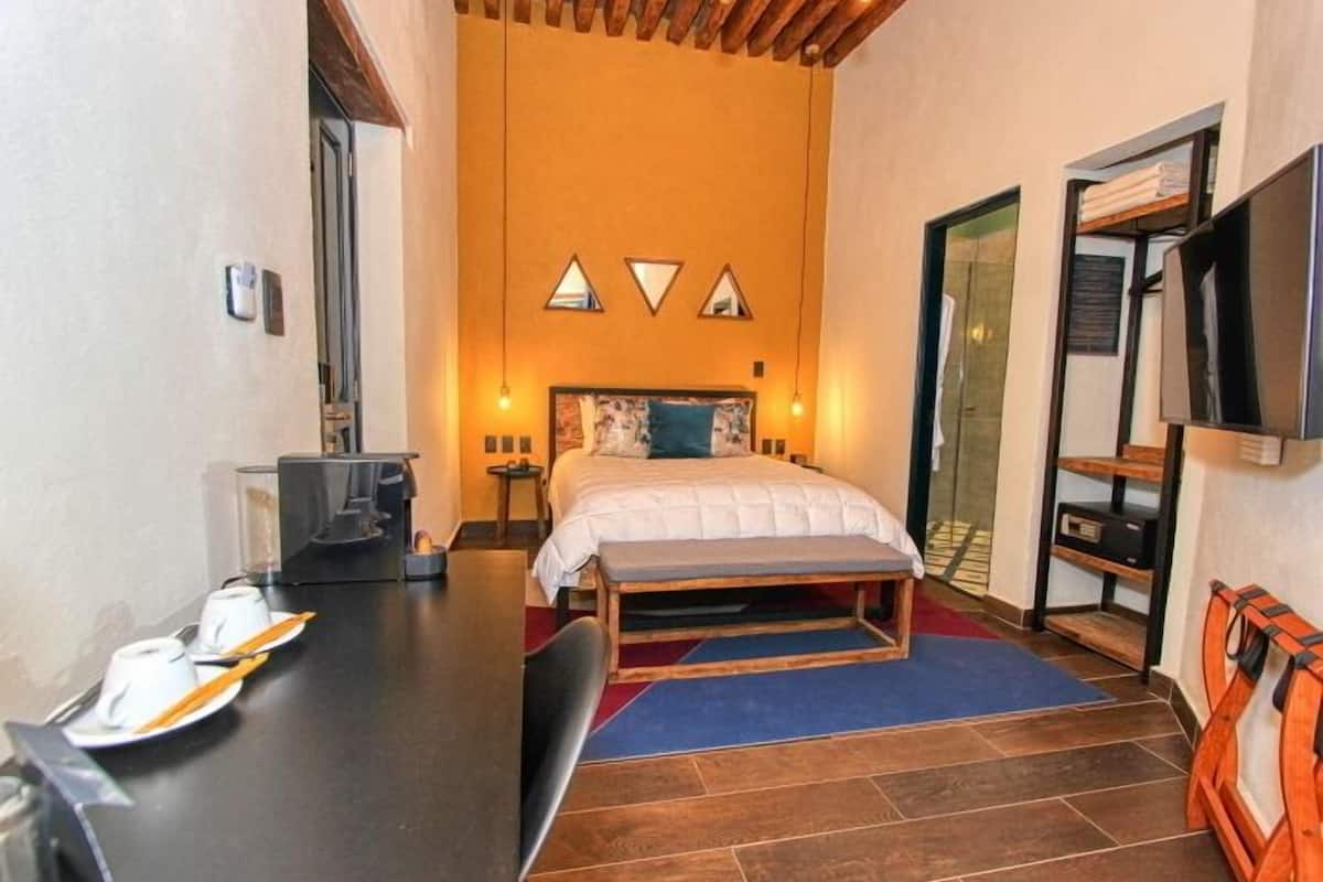 A compact hotel room with a bed, bedside tables, a desk with a chair, a bench, a TV, and a small shelf.