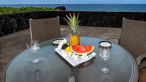 Ett runt glasbord med två stolar, en ananas, en skiva vattenmelon och två vinglas på en terrass med utsikt över havet.