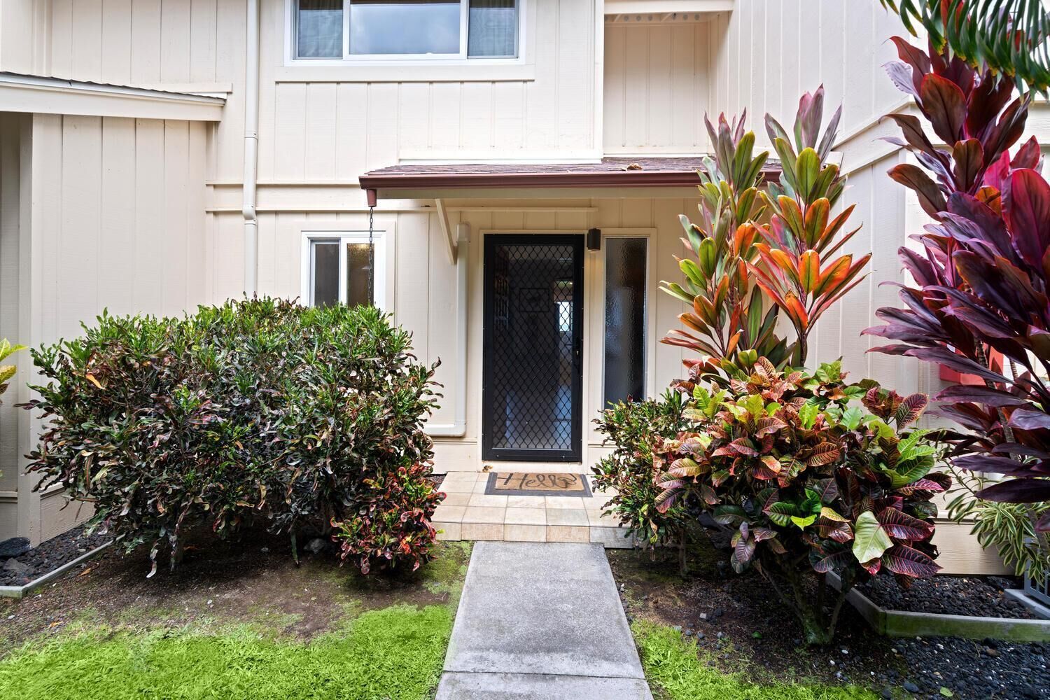 Um edifício residencial com porta frontal, um pequeno jardim com diversas plantas e um caminho de concreto que leva à porta.