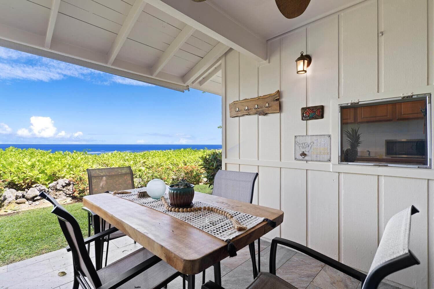 A covered patio with a wooden table and chairs, a view of the ocean, and a kitchen window with a pineapple.