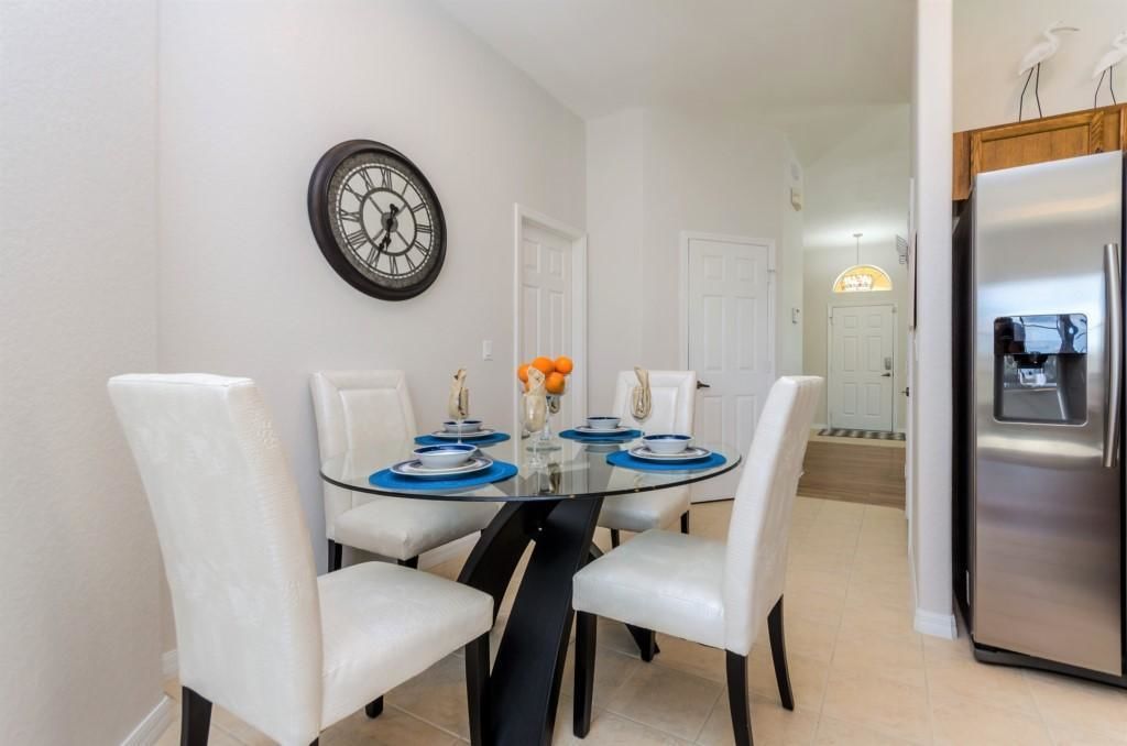 Un coin repas avec une table en verre, des chaises blanches, une horloge au mur et un réfrigérateur à l’arrière-plan.
