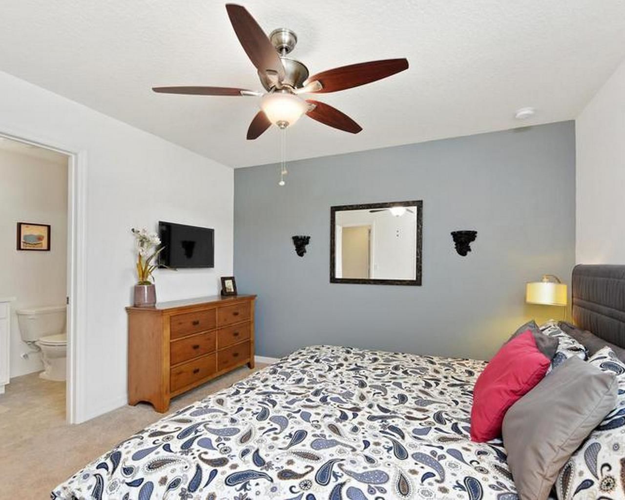 A bedroom with a bed, a ceiling fan, a dresser, a mirror, and a nightstand with a lamp.
