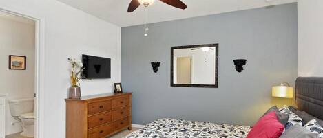 A bedroom with a bed, a ceiling fan, a dresser, a mirror, and a nightstand with a lamp.