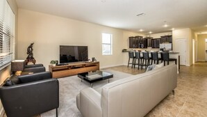 A modern living room with a sofa, TV, and entertainment unit. A kitchen area with bar stools is visible in the background.