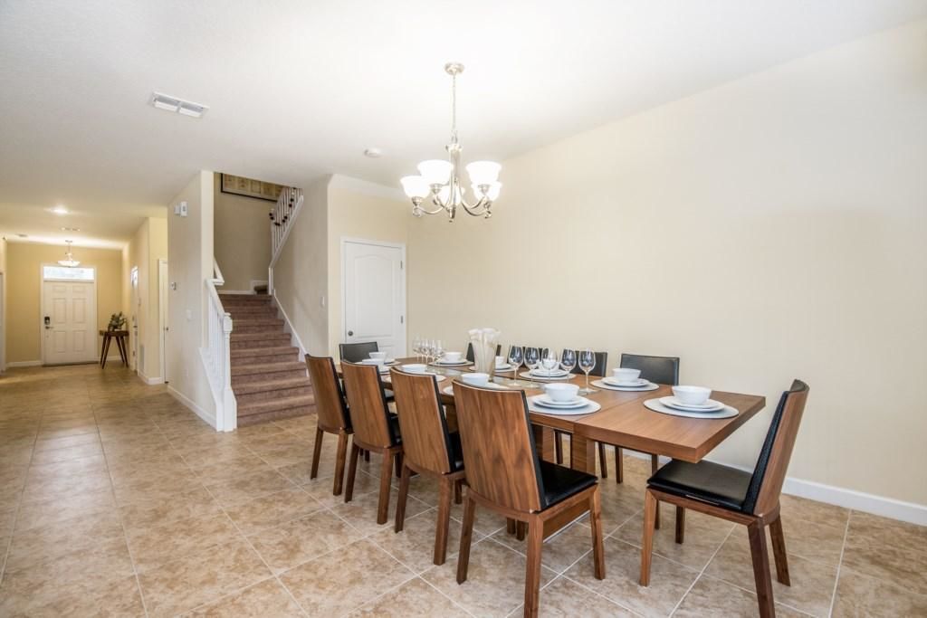 Un coin repas avec une table en bois dressée pour six personnes, entourée de six chaises dotées de coussins noirs. Un lustre est suspendu au plafond, et un escalier est visible au fond.