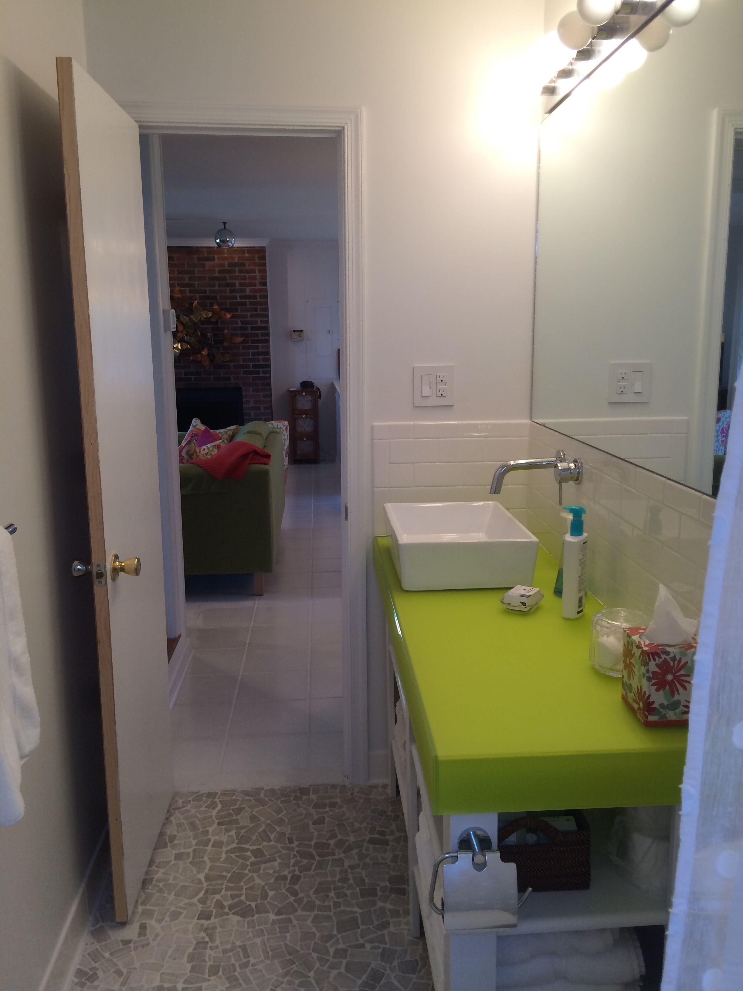 Un baño con encimera verde, lavabo blanco y espejo encima. Se ve una puerta que da a un área de estar con chimenea de ladrillo.