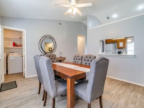 A dining area with a wooden table and grey upholstered chairs, a ceiling fan, and a kitchen area with a refrigerator and microwave.