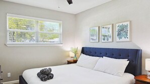 Ein Hotelzimmer mit einem großen Bett, einem Deckenventilator, einem Fenster mit Jalousien und drei gerahmten Kunstwerken an der Wand.