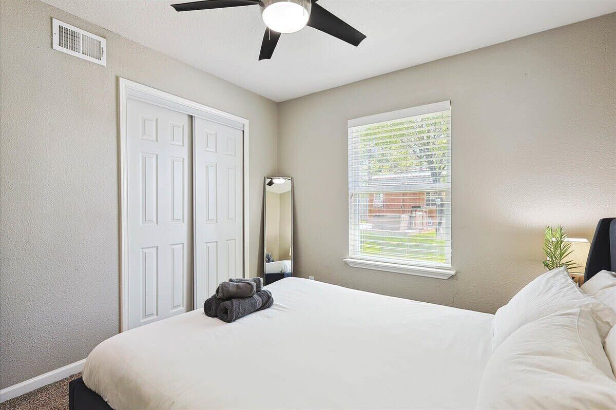 Une chambre à coucher comprenant un lit, un ventilateur de plafond, une fenêtre avec des stores, une porte et un miroir.