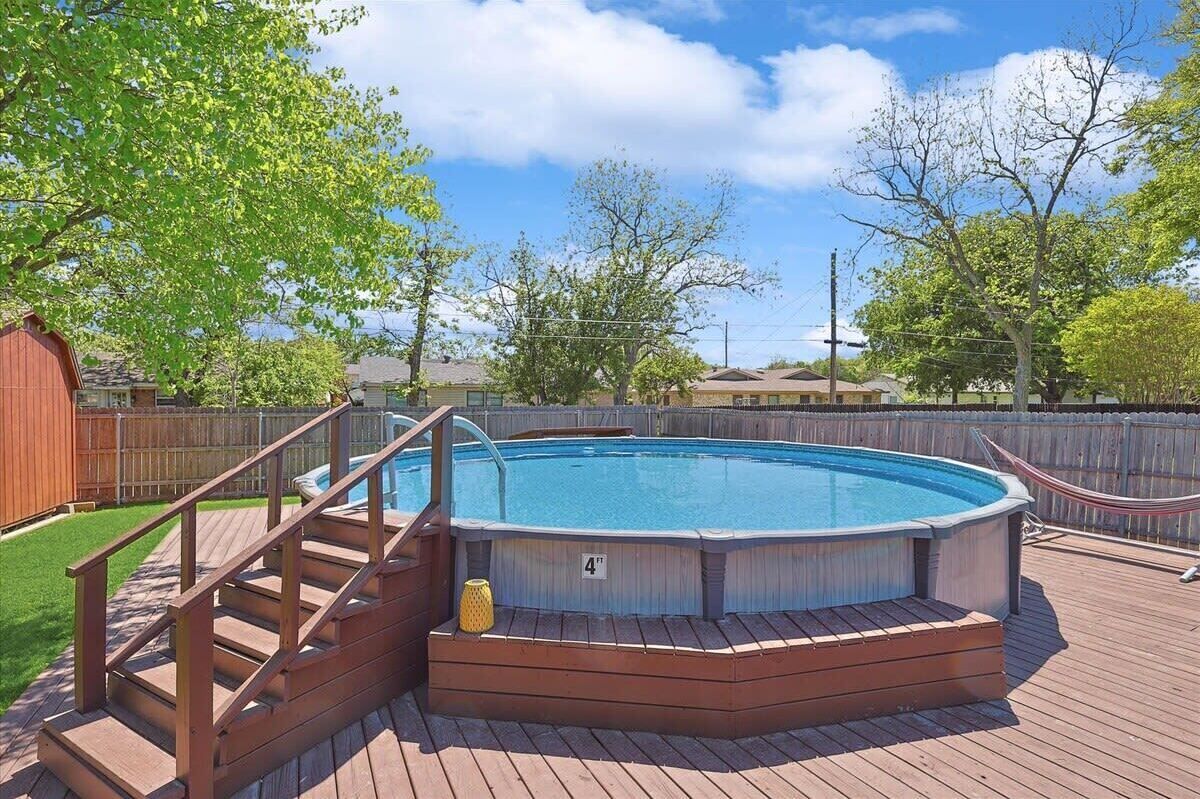 A rectangular above-ground pool with a wooden deck and stairs, surrounded by a wooden fence and trees.