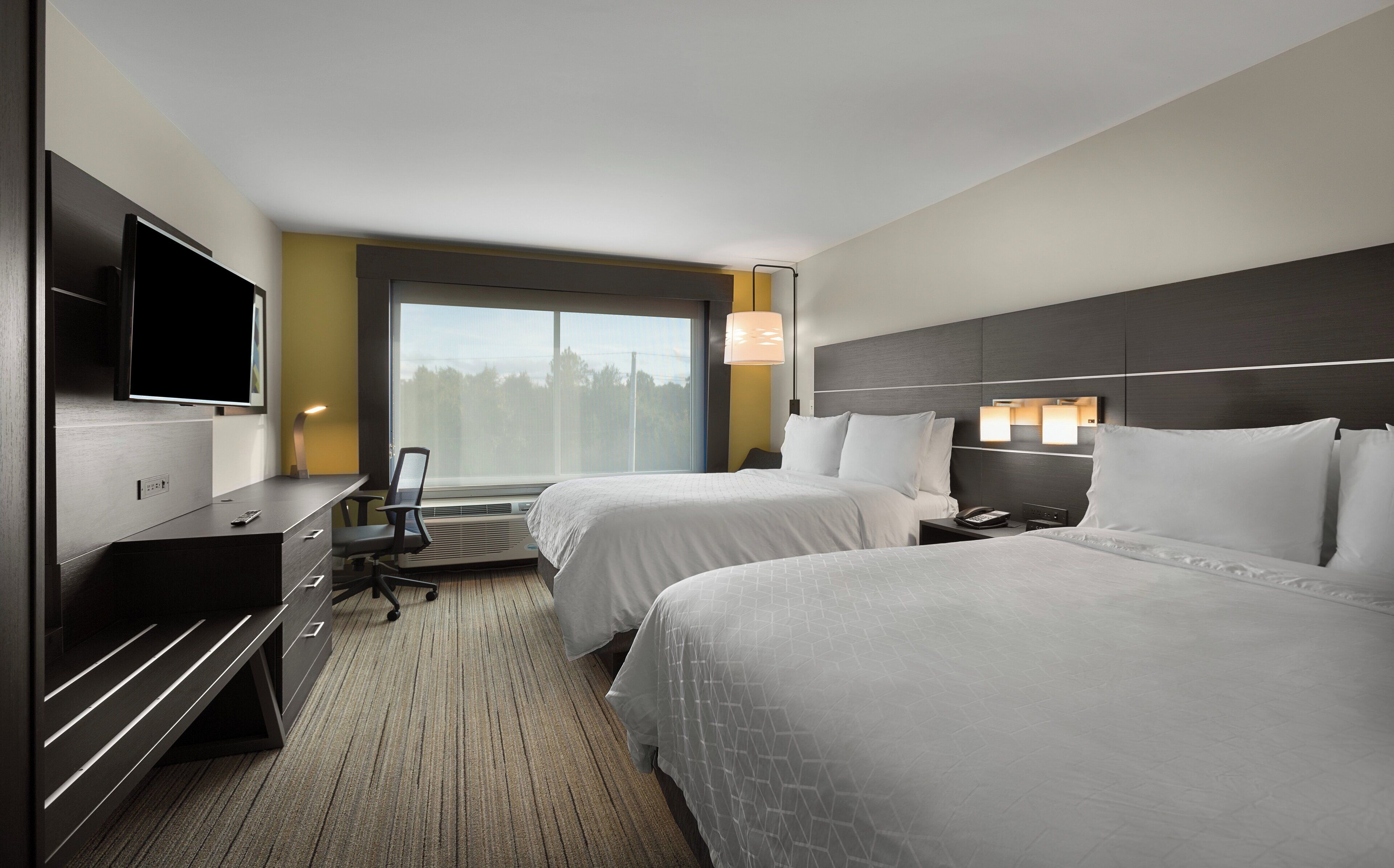 A hotel room with two beds, a desk, a TV, and a window with a view of trees.