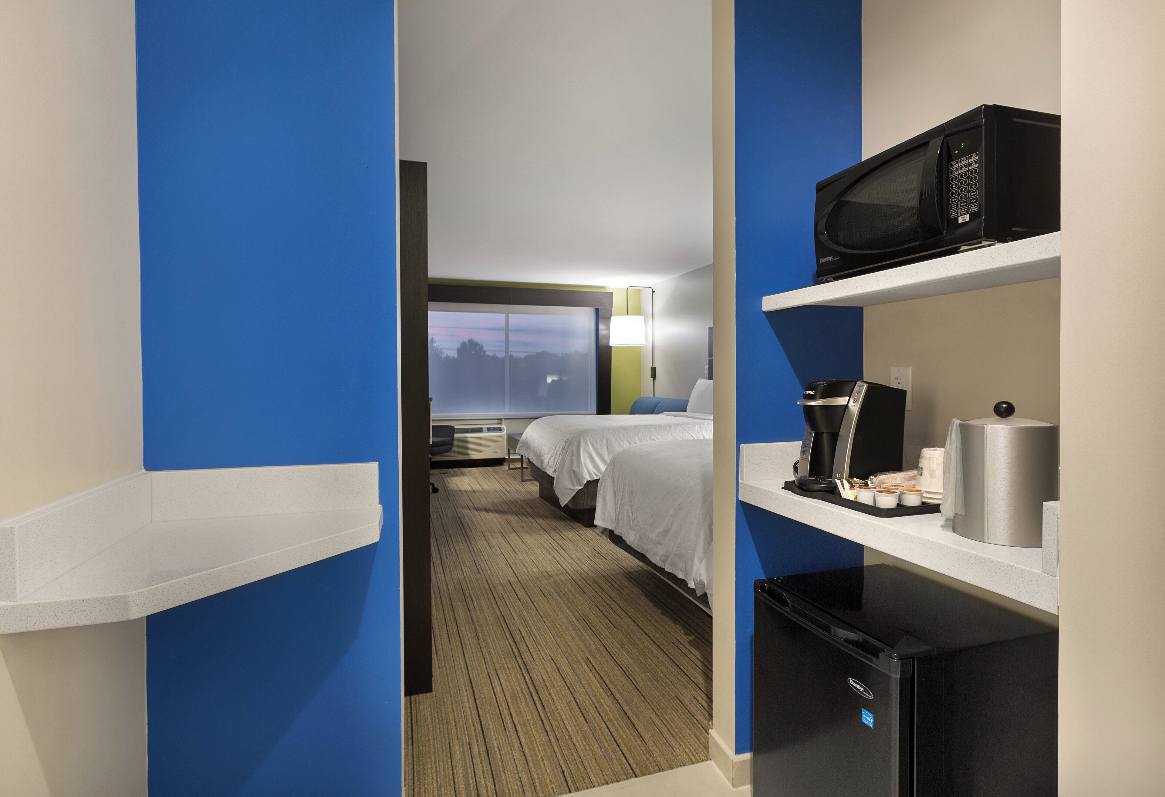 Habitación de hotel con cama, escritorio con microondas, cafetera y refrigerador pequeño.