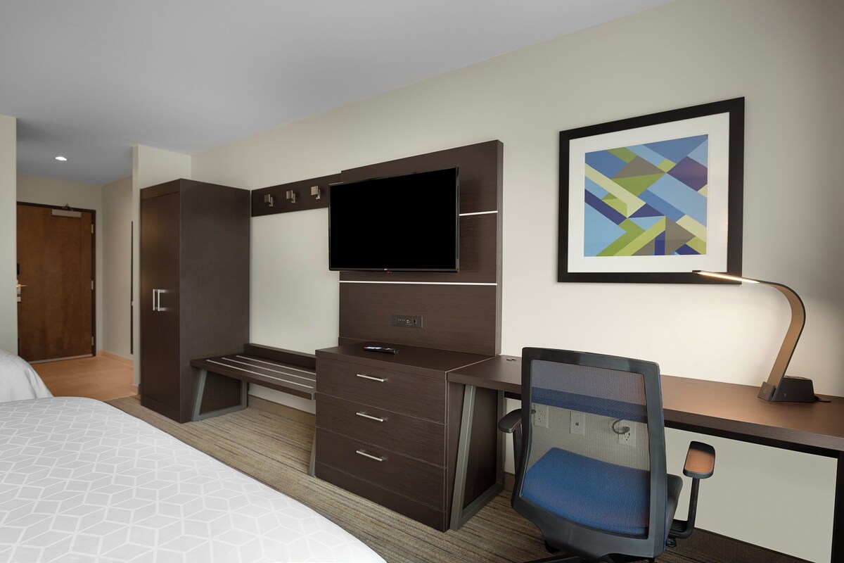 A hotel room with a bed, a desk with a chair, a TV, a wardrobe, and a framed picture on the wall.
