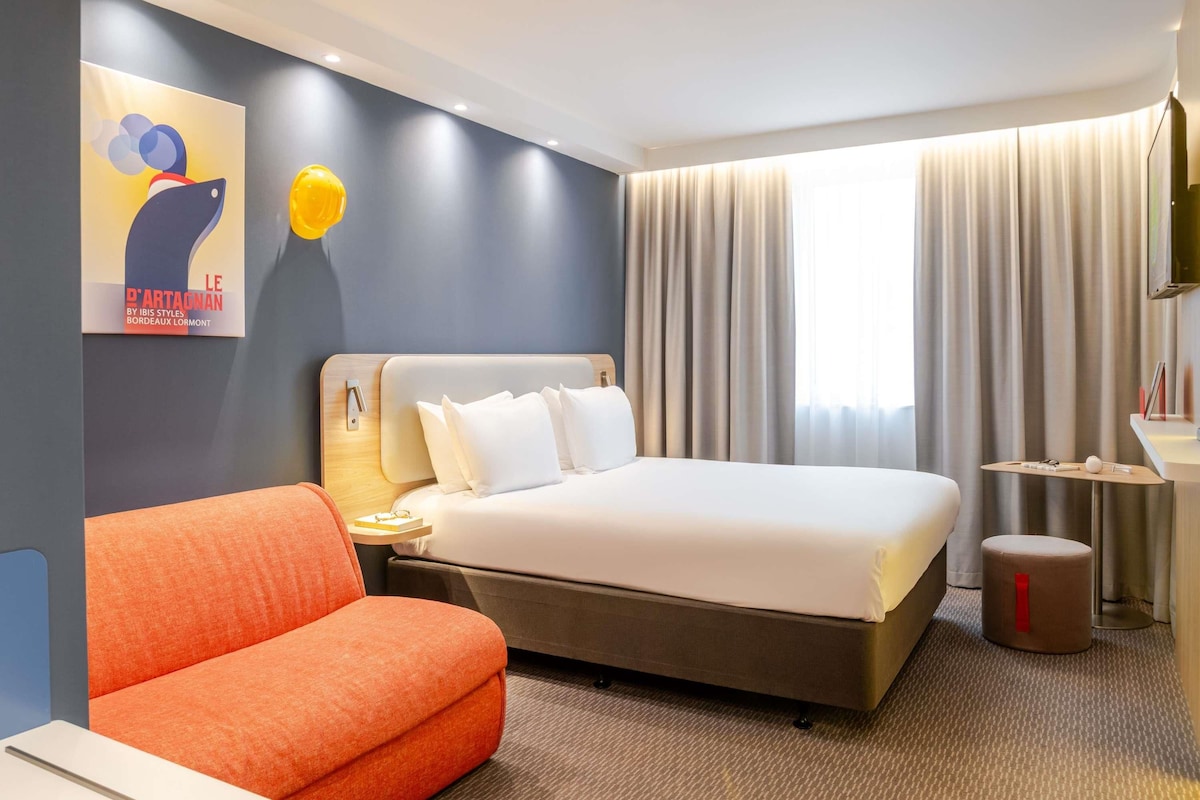 A hotel room with a bed, a nightstand, a chair, a small table, and a television.
