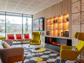 A modern living room with a fireplace, a television mounted on the wall, and a large window offering a view of the city. - ibis Styles Bordeaux Lormont (Lormont)