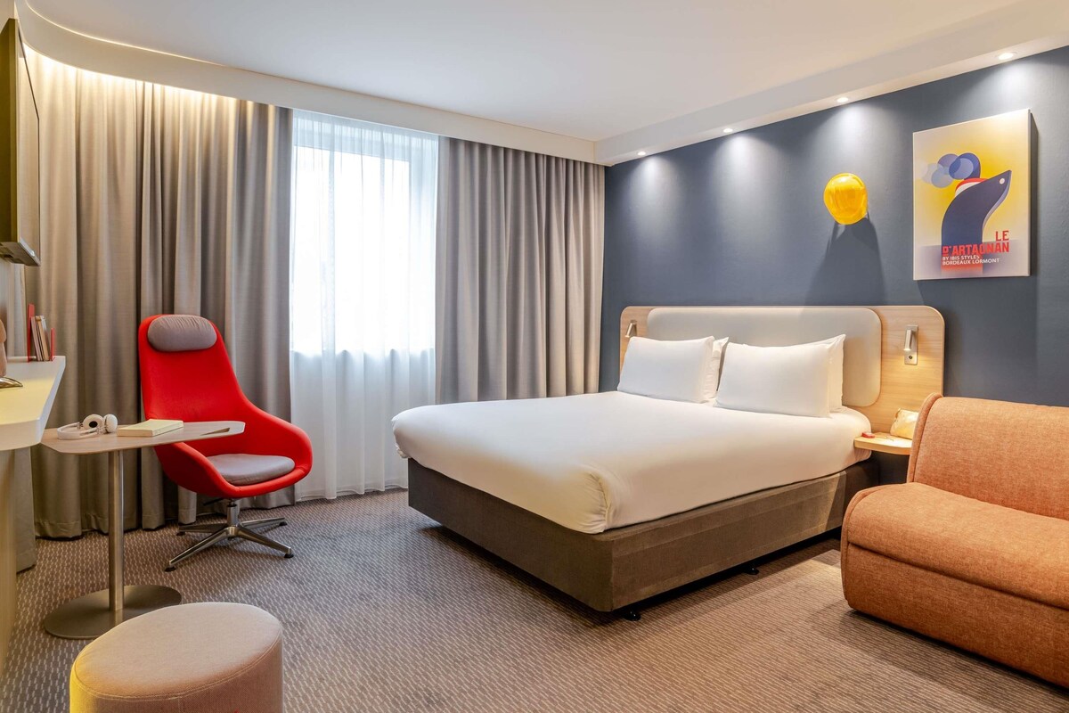 A hotel room with a bed, a red chair, a small table, and a painting on the wall.