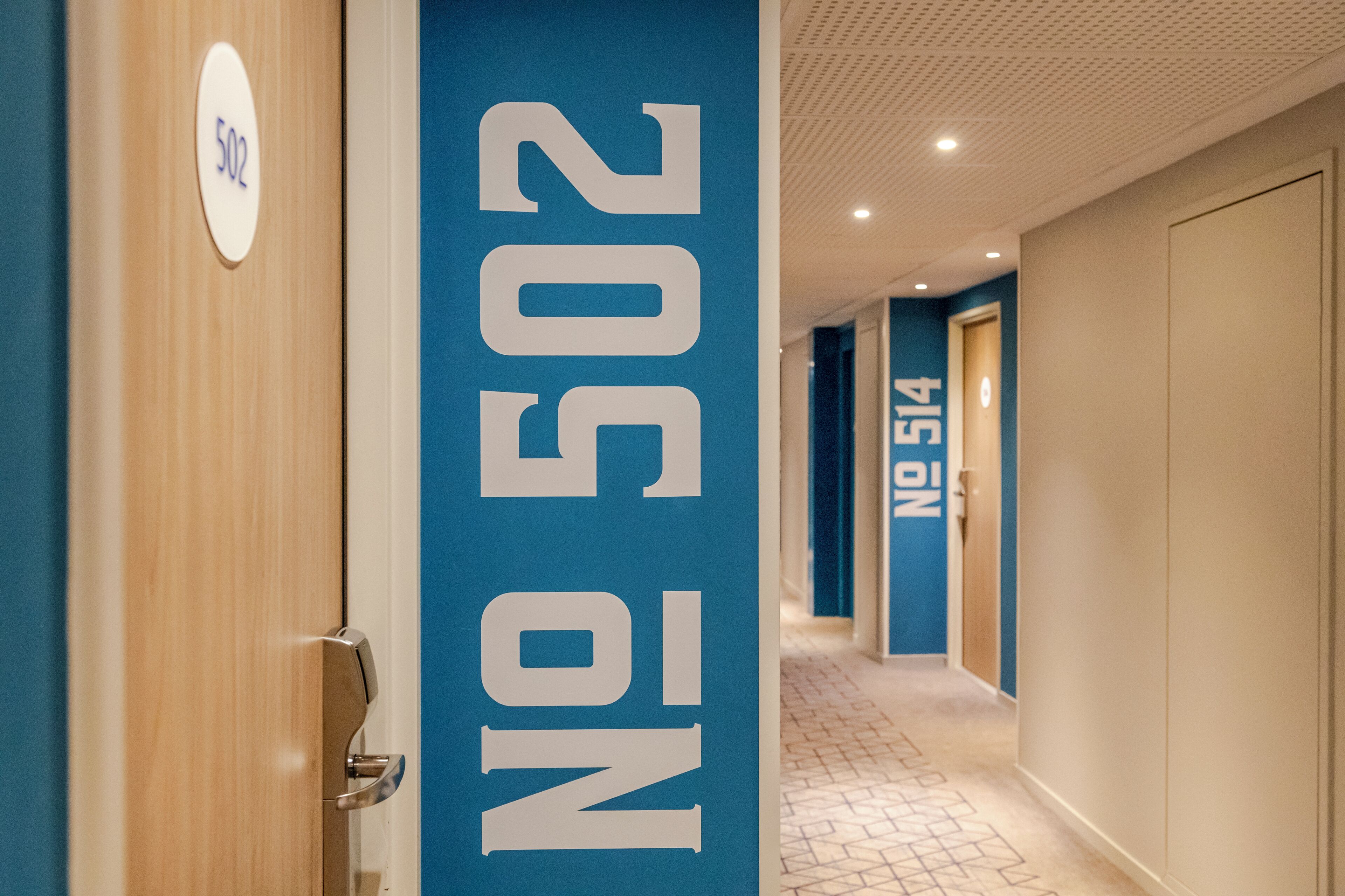 Un couloir d’hôtel aux murs bleus, avec les portes numérotées 502 et 514.