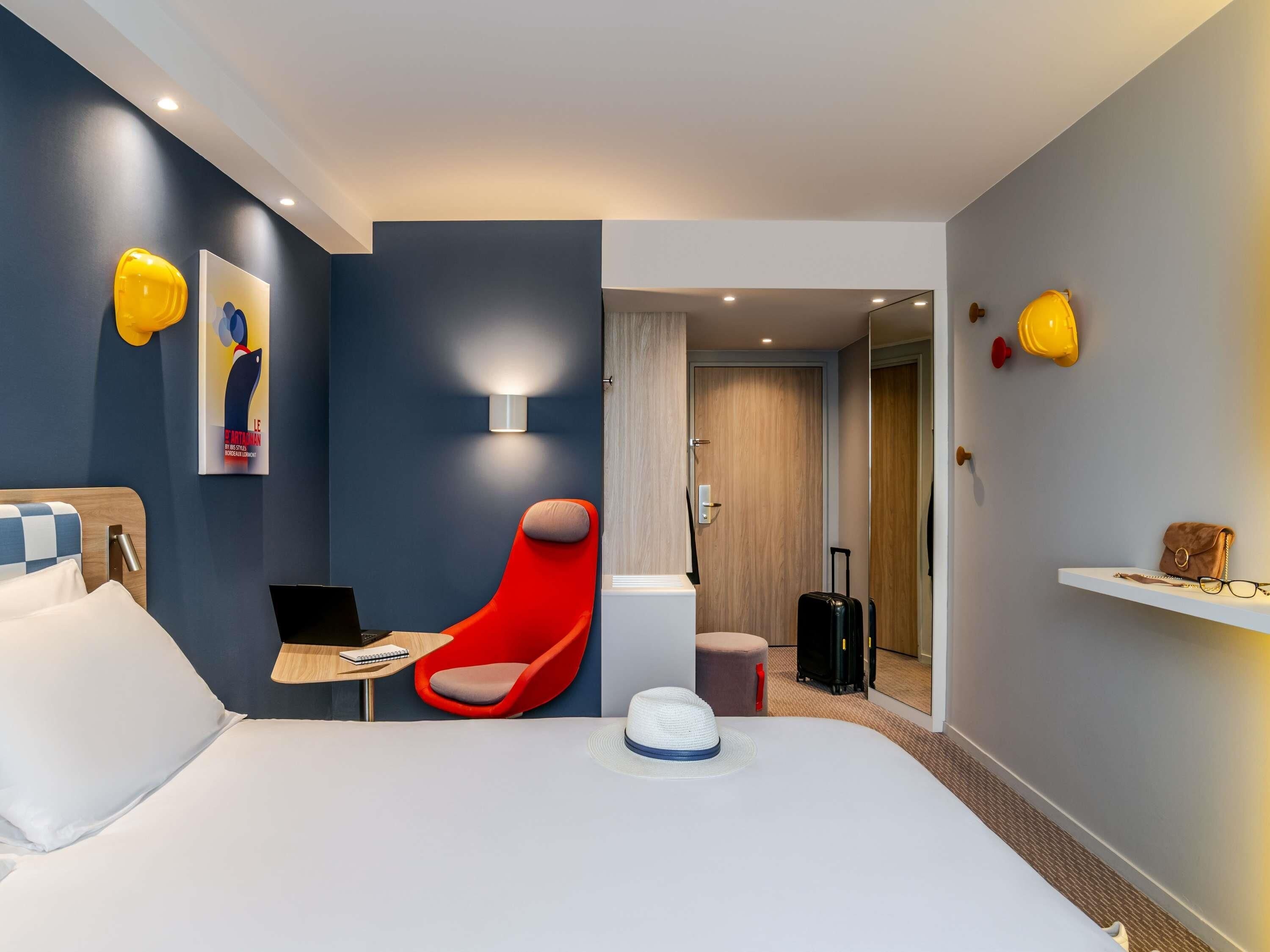 Une chambre d’hôtel avec un lit, un bureau avec un ordinateur portable, une chaise rouge, une valise et une étagère murale sur laquelle est posé un sac à main.