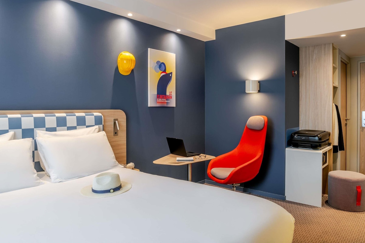 A hotel room with a bed, a desk, a chair, a suitcase, and a wall-mounted lamp.