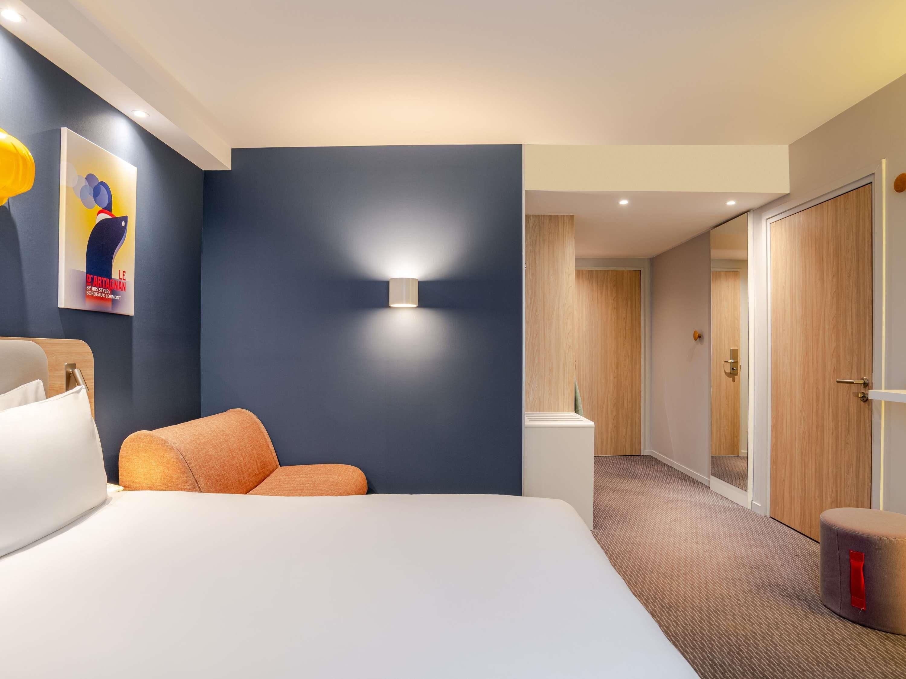 A hotel room with a large bed, a bedside table, a wall-mounted light, a poster, and a door leading to another room.