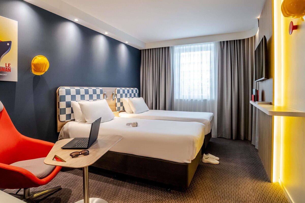 A hotel room with two beds, a desk with a laptop, a chair, and a window with curtains.