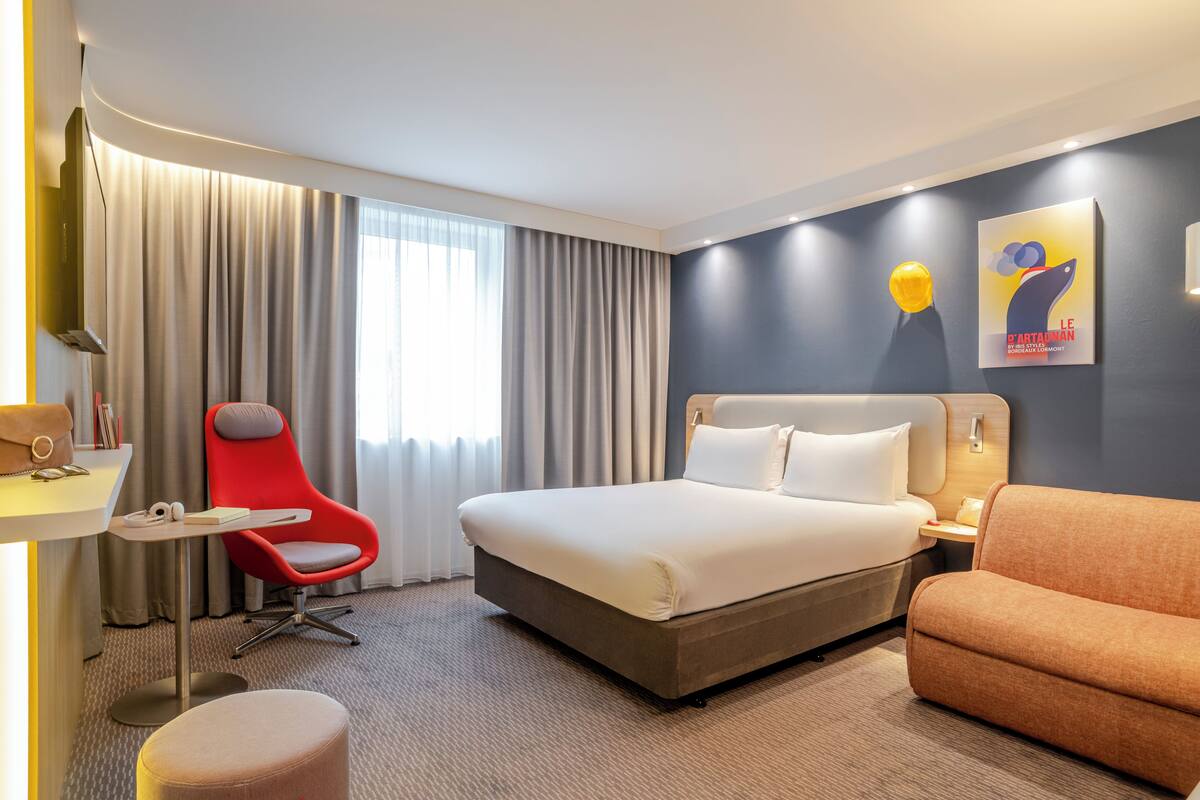 Une chambre d’hôtel avec un grand lit, un fauteuil rouge, un canapé orange, une petite table et une grande fenêtre avec des rideaux.