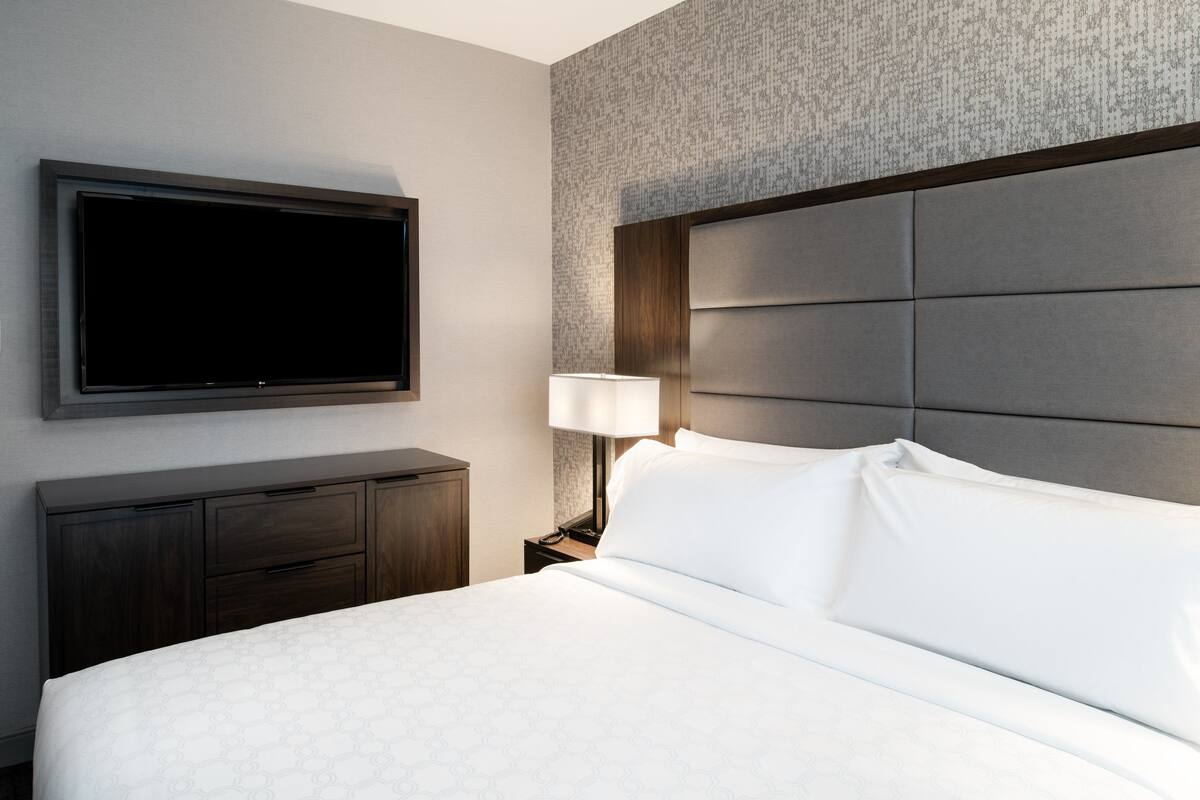 A hotel room with a large bed, a flat-screen TV mounted on the wall, a bedside table with a lamp, and a dark wooden dresser.