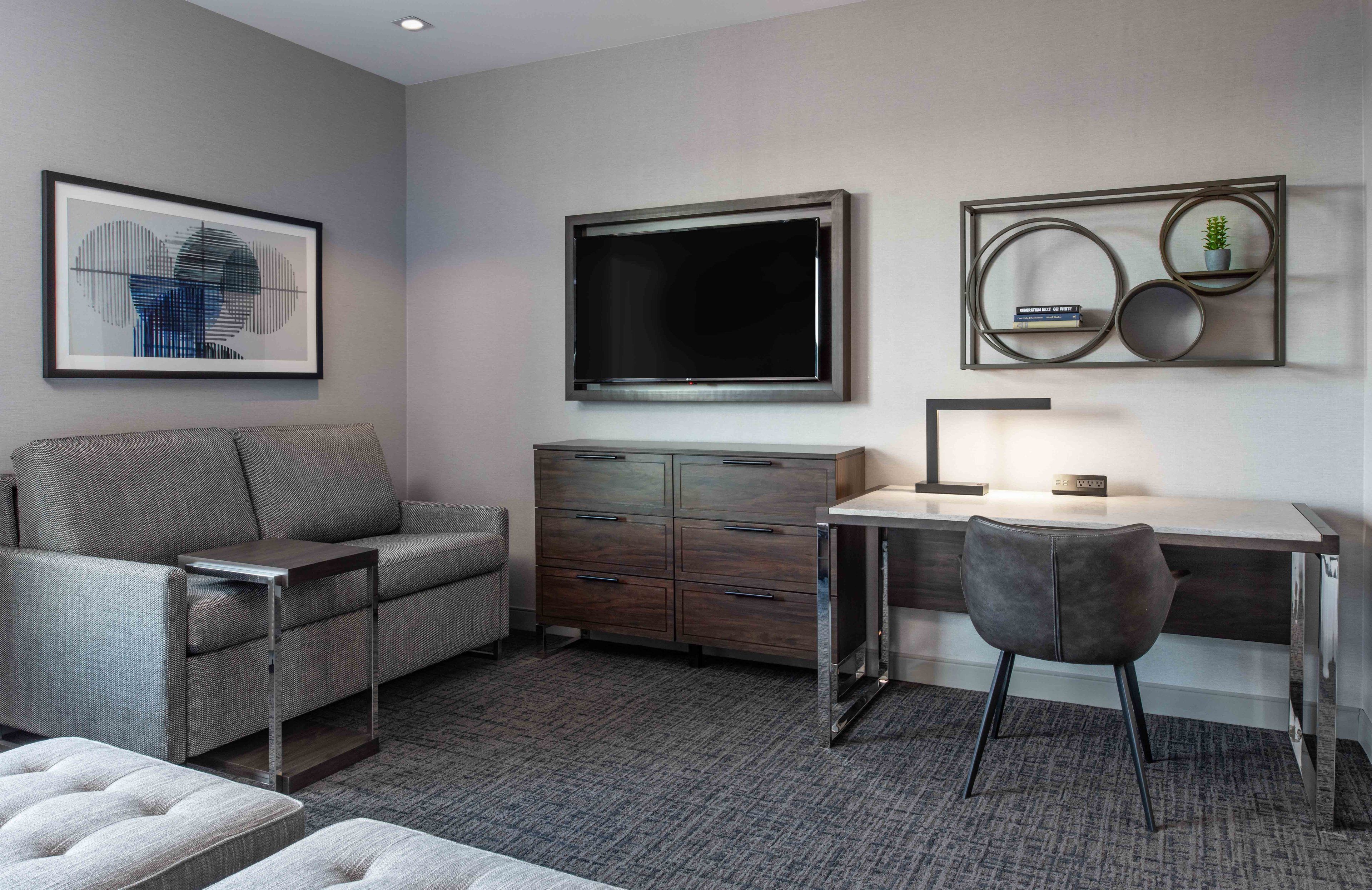 A modern living room with a grey sofa, a wooden cabinet, a desk with a chair, and a large TV mounted on the wall.