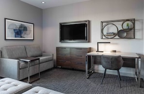 A modern living room with a grey sofa, a wooden cabinet, a desk with a chair, and a large TV mounted on the wall. - Staybridge Suites Boston - Quincy by IHG (Quincy)