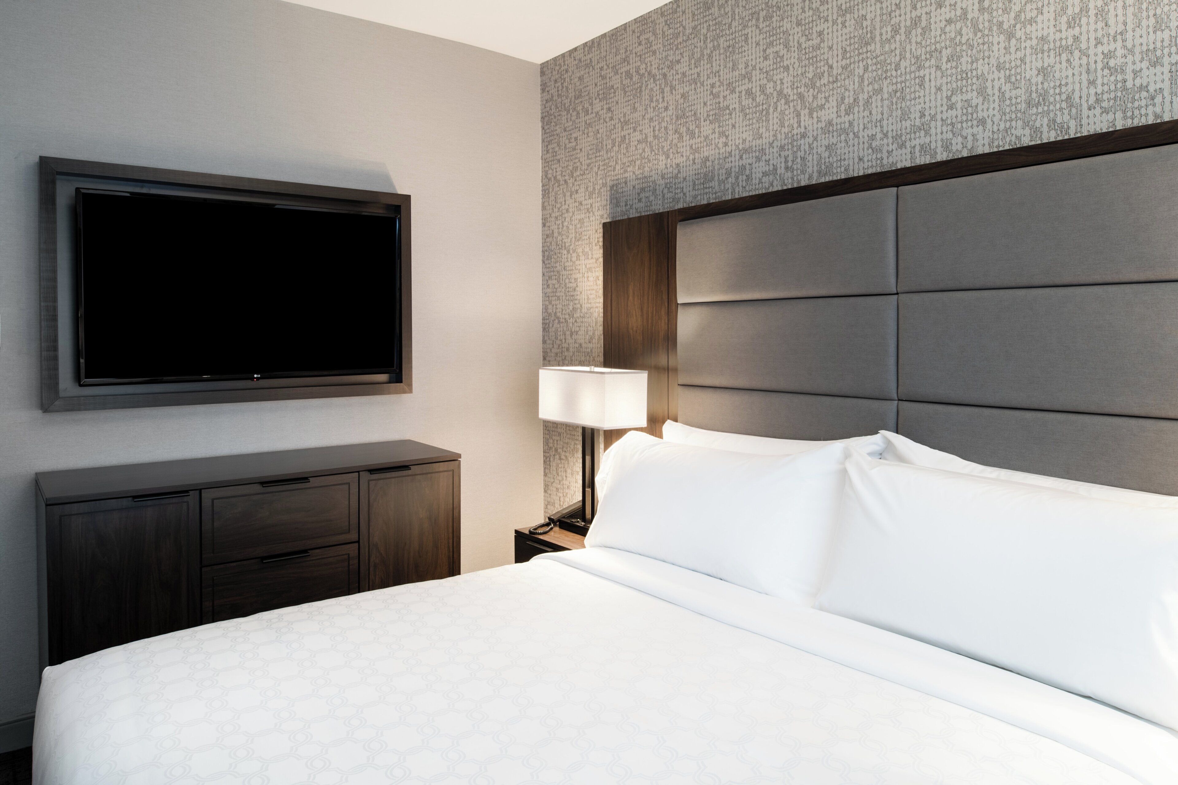 Una habitación de hotel con una cama grande, un televisor de pantalla plana montado en la pared, una mesita de noche con una lámpara y un aparador de madera oscura.