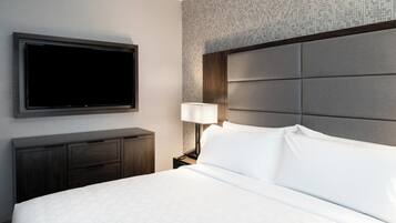Una habitación de hotel con una cama grande, un televisor de pantalla plana montado en la pared, una mesita de noche con una lámpara y un aparador de madera oscura.