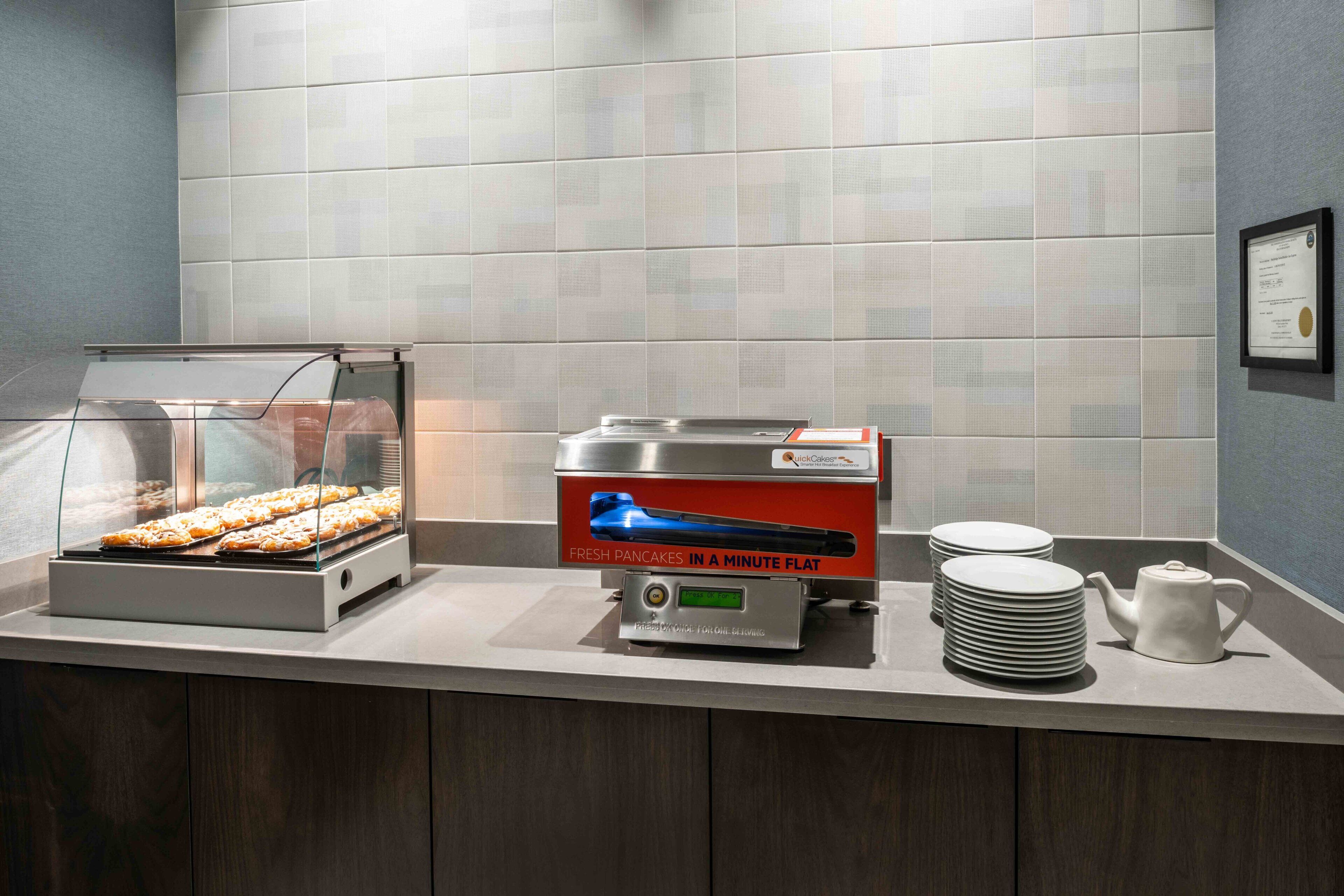 A breakfast station with a display case of pastries, a pancake maker, and stacked plates and a teapot.