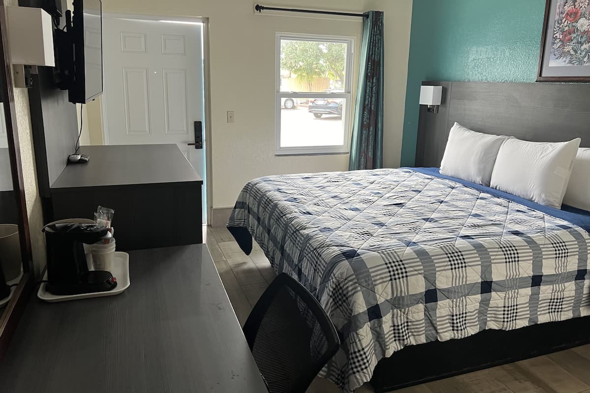 A hotel room with a bed, a desk with a computer, a chair, a window with a view of the outside, and a door.