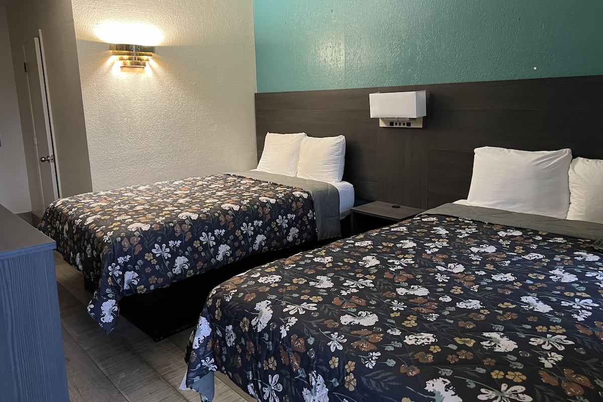 A hotel room with two beds, each with floral-patterned bedding, a nightstand, a wall-mounted light, and a door.