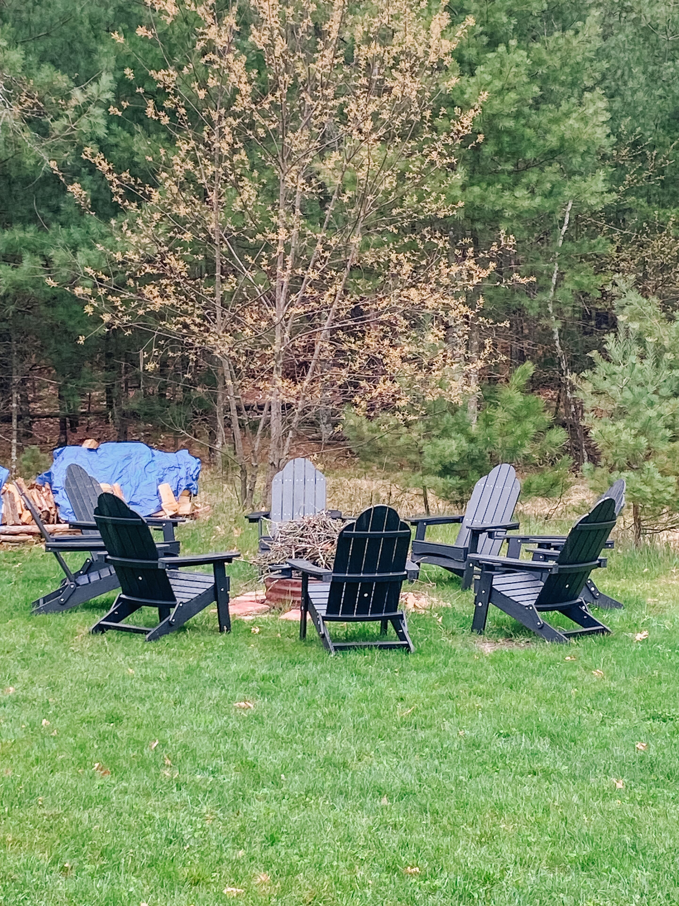 Vier Adirondack-Stühle sind um eine Feuerstelle in einer grünen Außenanlage angeordnet, im Hintergrund sind Bäume zu sehen.
