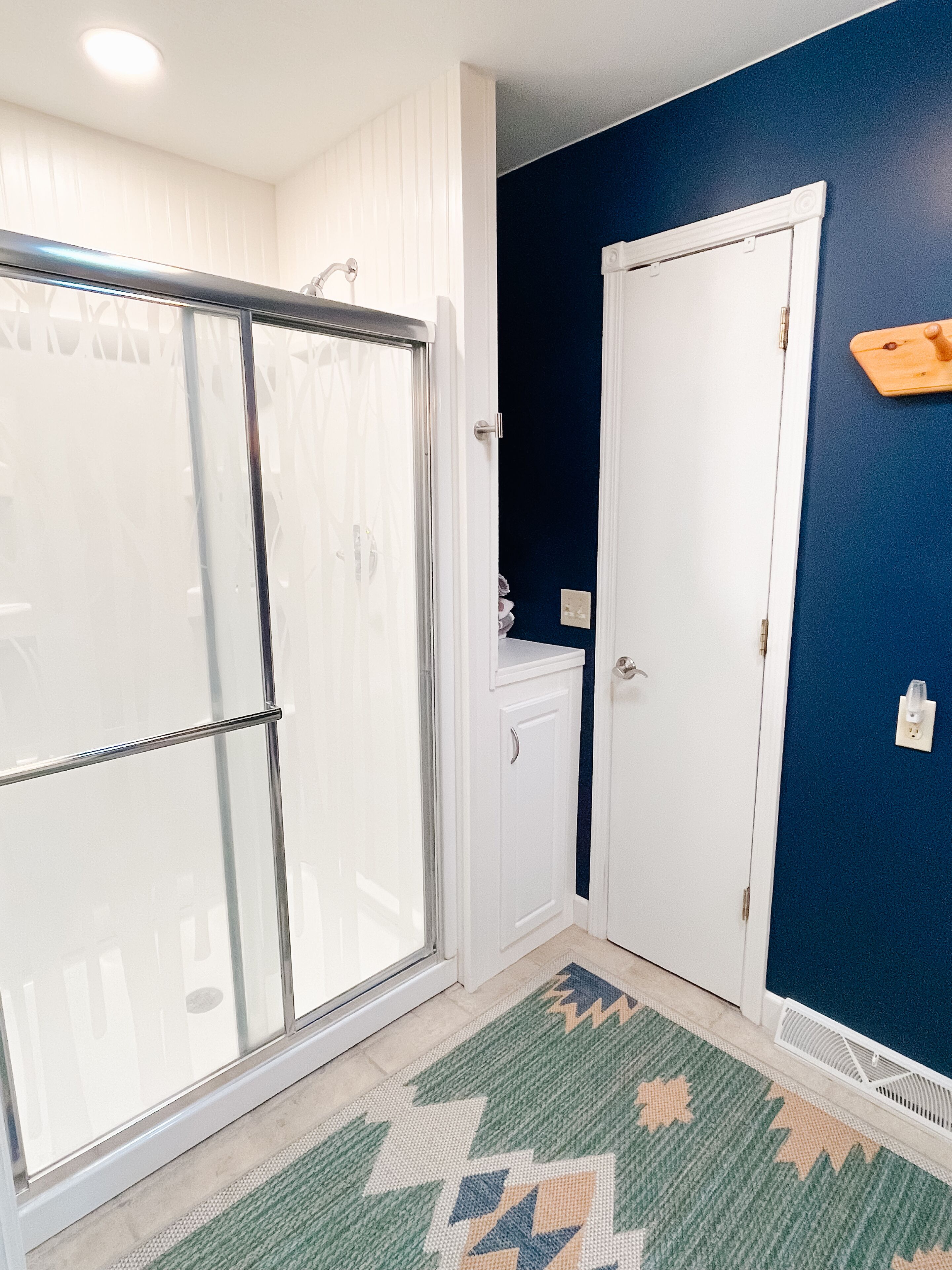A bathroom with a glass shower door, a white cabinet, a blue wall, and a patterned rug.