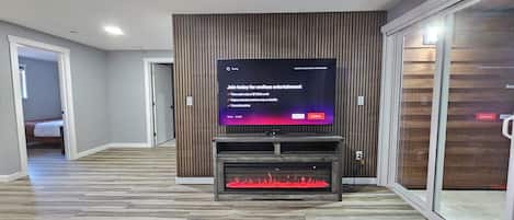 Una sala de estar moderna con un televisor de pantalla plana montado en una pared de madera, una chimenea debajo y un ventanal de vidrio a la derecha.