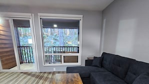 A modern living room with a dark grey sectional sofa, a wooden coffee table, and a large sliding glass door leading to a balcony with a view of trees. - Olympic Suites Inn (Forks)